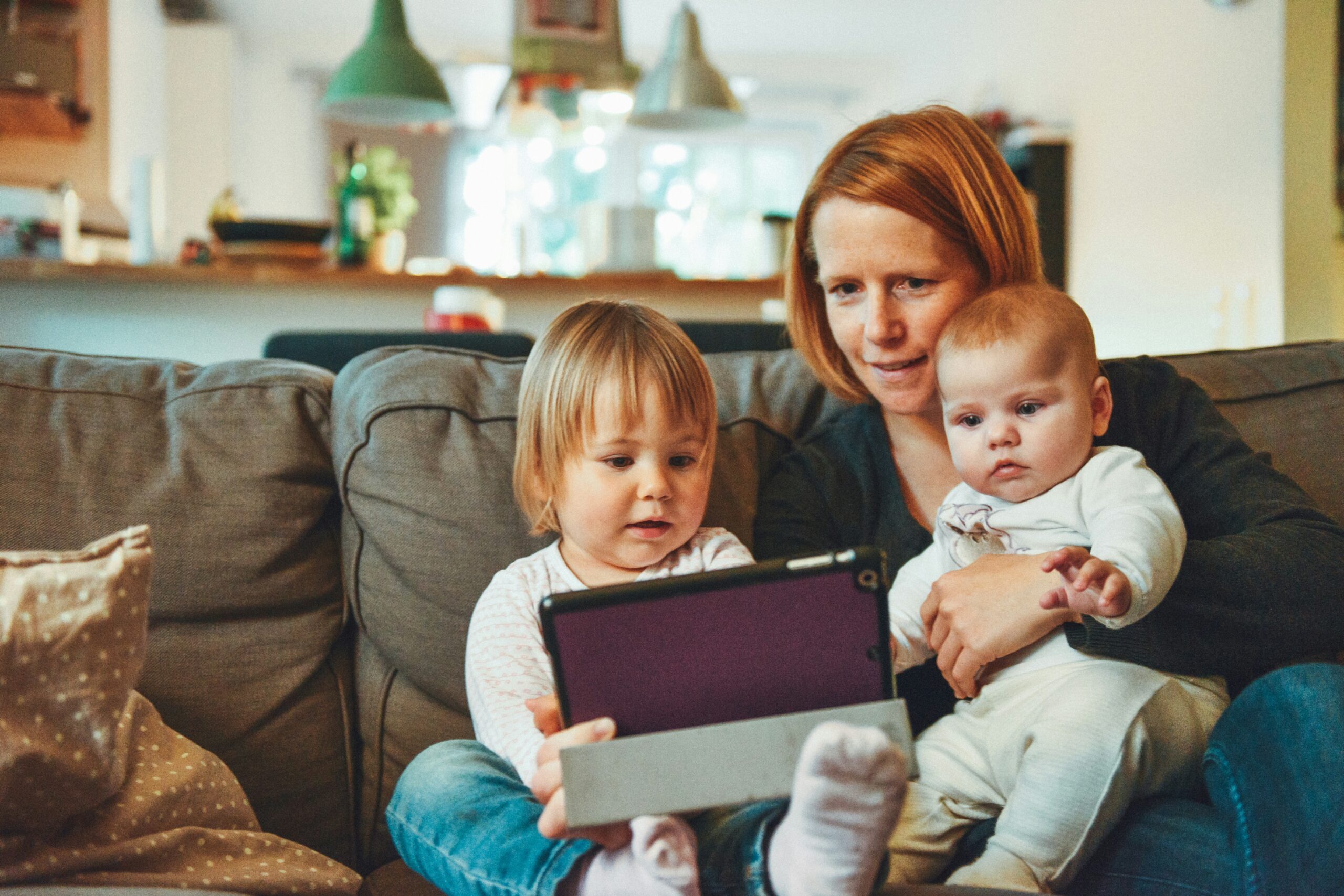 Mother snuggling a baby and toddler on the couch watching a tablet computer.